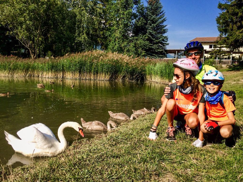Famiglia in vacanza con Girolibero accanto a cigni in un laghetto alpino nei dintorni di Bolzano.