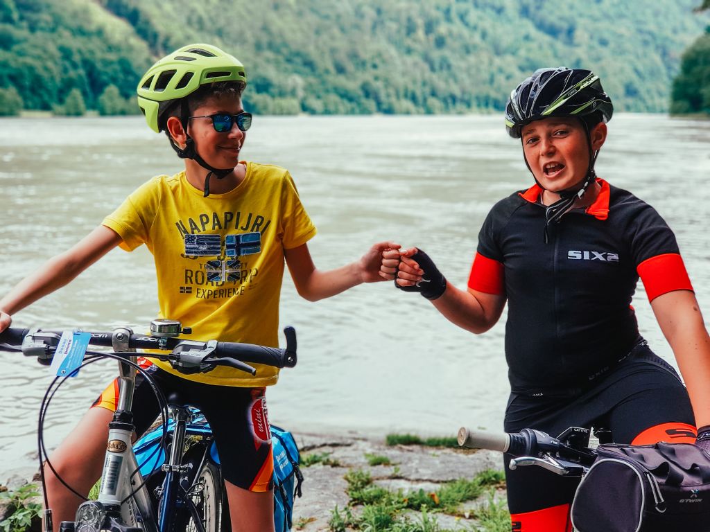 Radausflüge mit der Familie, zwei kleine Radfahrer, die sich am Fluss die Hände reichen, mit einer grünen Landschaft im Hintergrund