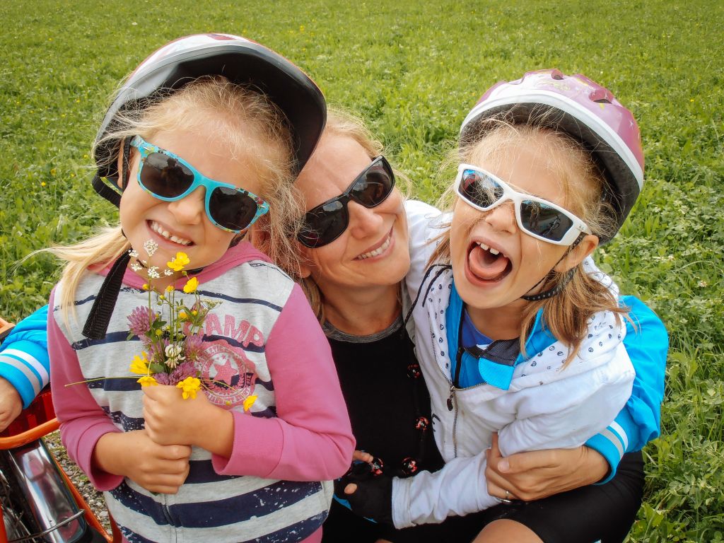 Mamma abbracciata con bambine sorridenti su un prato fiorito, vacanza all'aria aperta in bicicletta, ciclabile della Drava, Austria