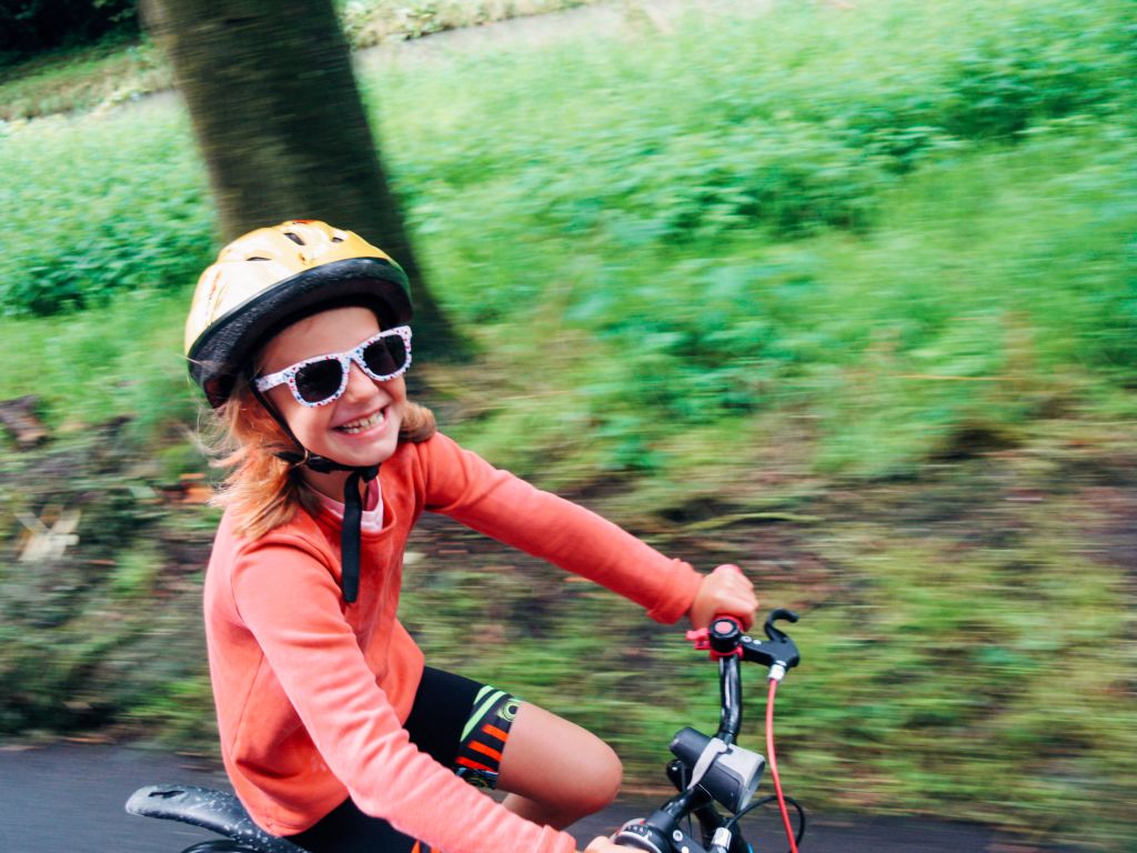 Bambina sorridente in bici, occhiali da sole, caschetto, tronco di un albero, prato verde, Belgio