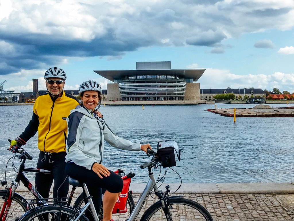 Coppia di ciclisti in viaggio con Girolibero lungo la ciclabile da Berlino a Copenaghen in posa per una foto davanti a un canale del Nord con edificio moderno sullo sfondo