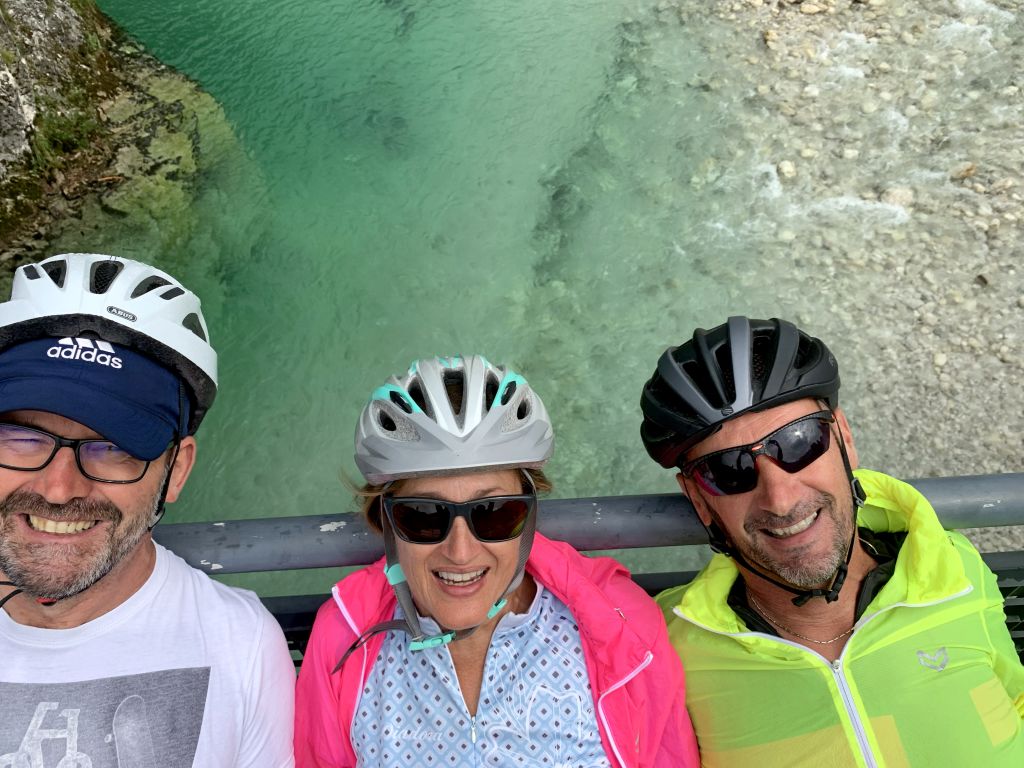 Ciclisti in posa vicino a un fiume verde smeraldo lungo la Ciclovia Alpe Adria, con casco e abbigliamento sportivo.