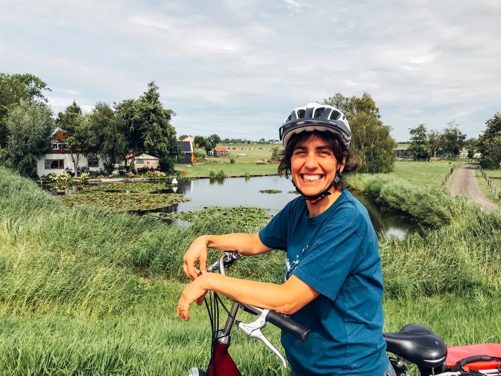 Donna sorridente in bicicletta in viaggio con Girolibero tra i campi verdi della campagna olandese, perfetta per il cicloturismo.