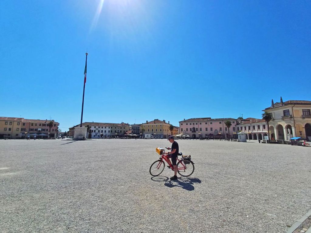 Cicloturista che attraversa la grande piazza di Grado, Italia, punto di arrivo della Ciclovia Alpe Adria