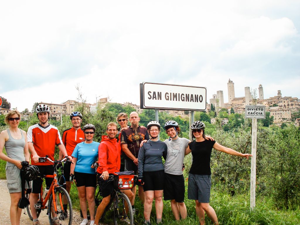 gruppo di ciclisti in posa davanti al cartello "San Gimignano", con centro storico della città sullo sfondo