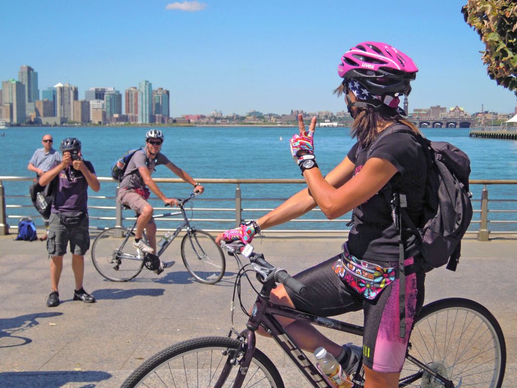 Ciclisti in viaggio con "Girolibero" che osservano la skyline di Manhattan lungo un percorso costiero.
