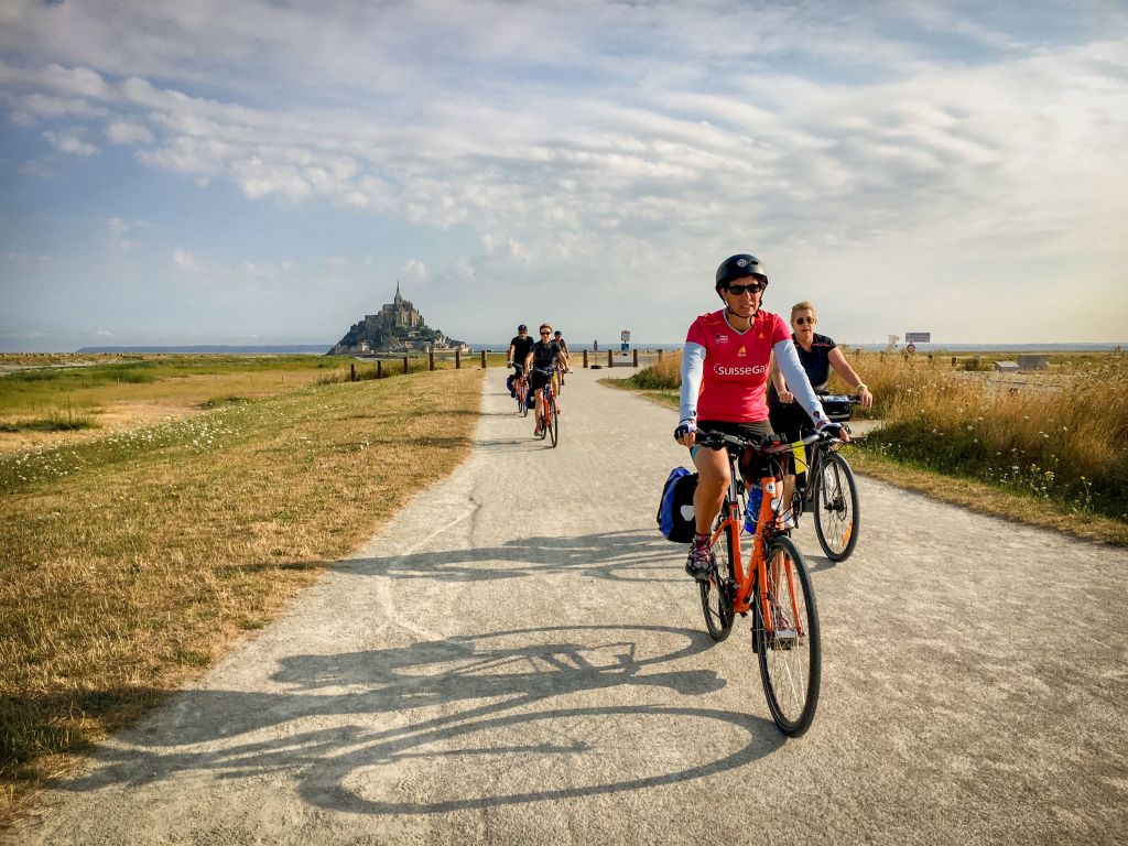 Ciclisti in vacanza con Girolibero su una strada panoramica verso Mont Saint-Michel, circondati da un paesaggio naturale.