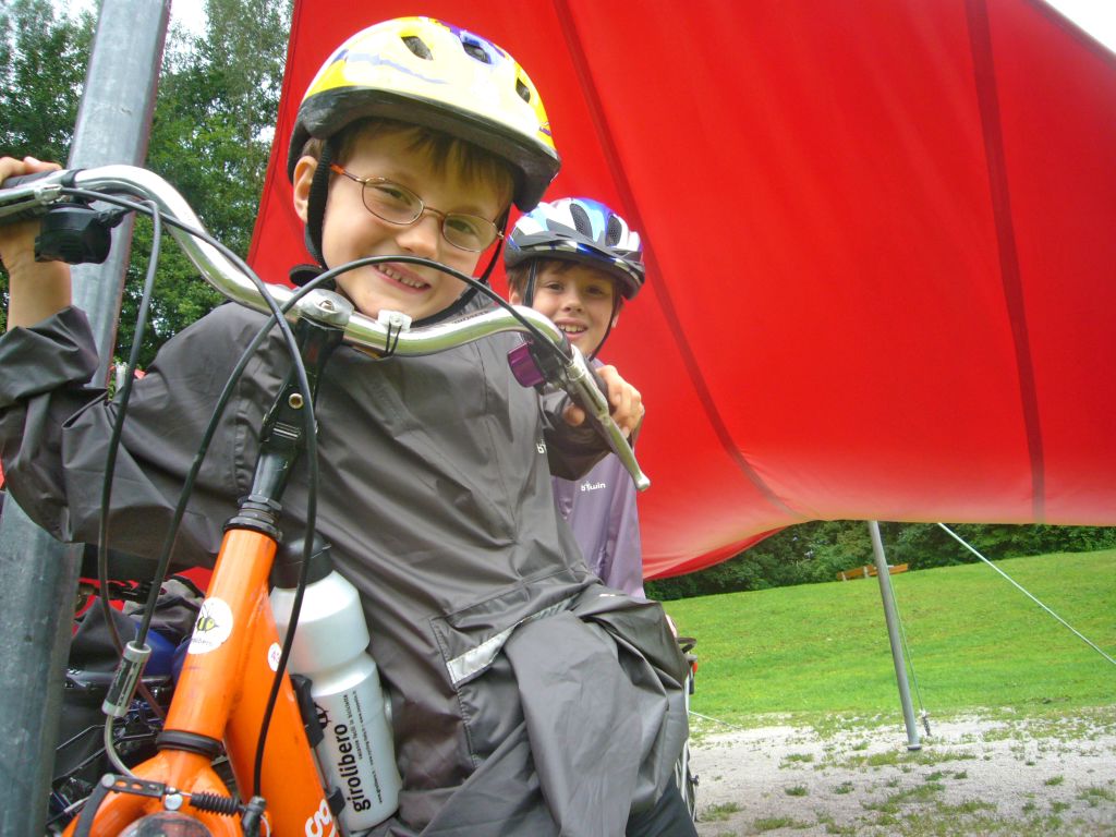 Bambini con casco e mantella impermeabile sotto un telo rosso, pronti per pedalare.