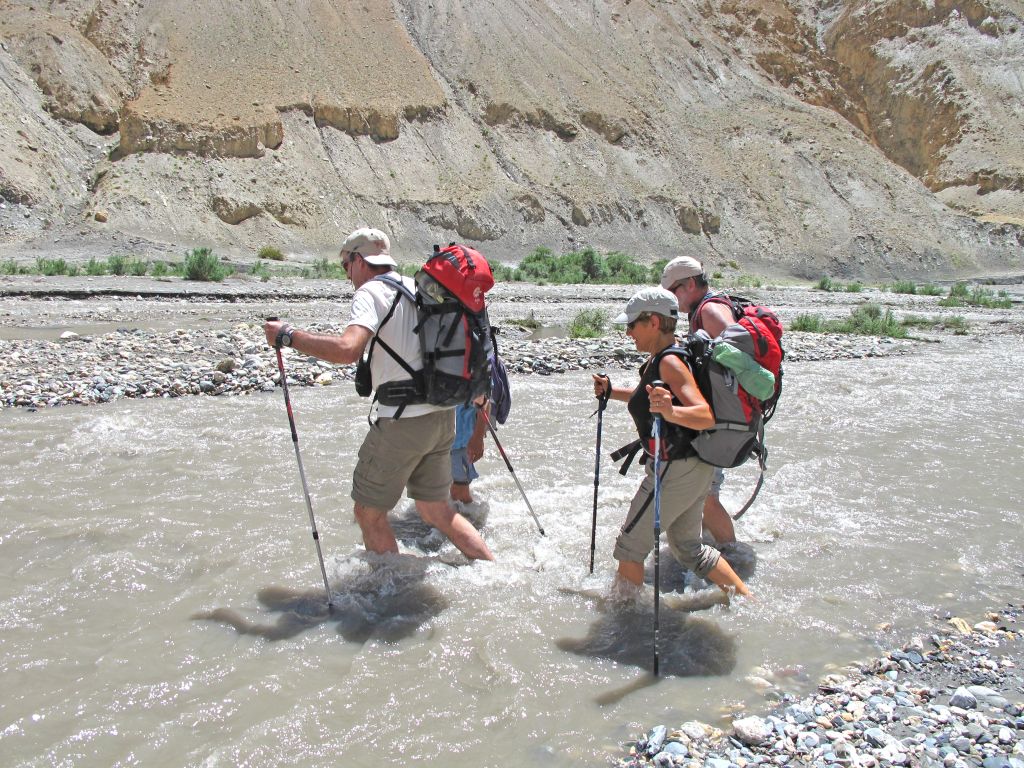 Escursionisti che attraversano un fiume con bastoni da trekking in un paesaggio montano del Ladakh.