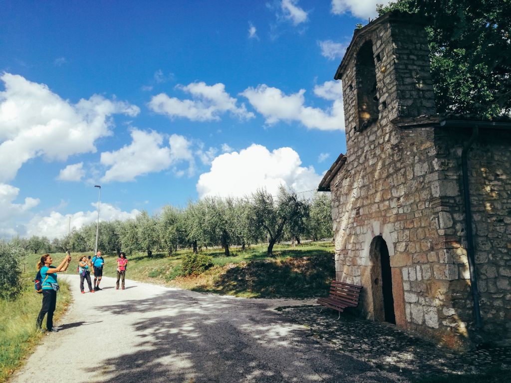 Strada alberata nella campagna umbra con chiesetta in pietra, colline verdi, escursionisti durante un trekking sulla Via di San Francesco