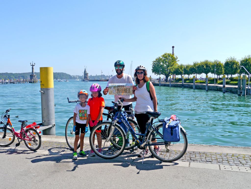 Famiglia che pedala lungo la pista ciclabile del Lago di Costanza con vista sull'acqua e porto turistico.