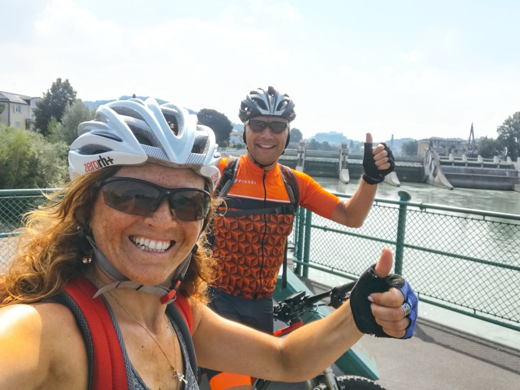 Coppia in bicicletta con casco e occhiali da sole lungo un fiume in Austria.