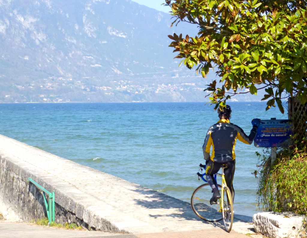 Ciclista che ammira il Lago di Ginevra da un muretto con montagne sullo sfondo.