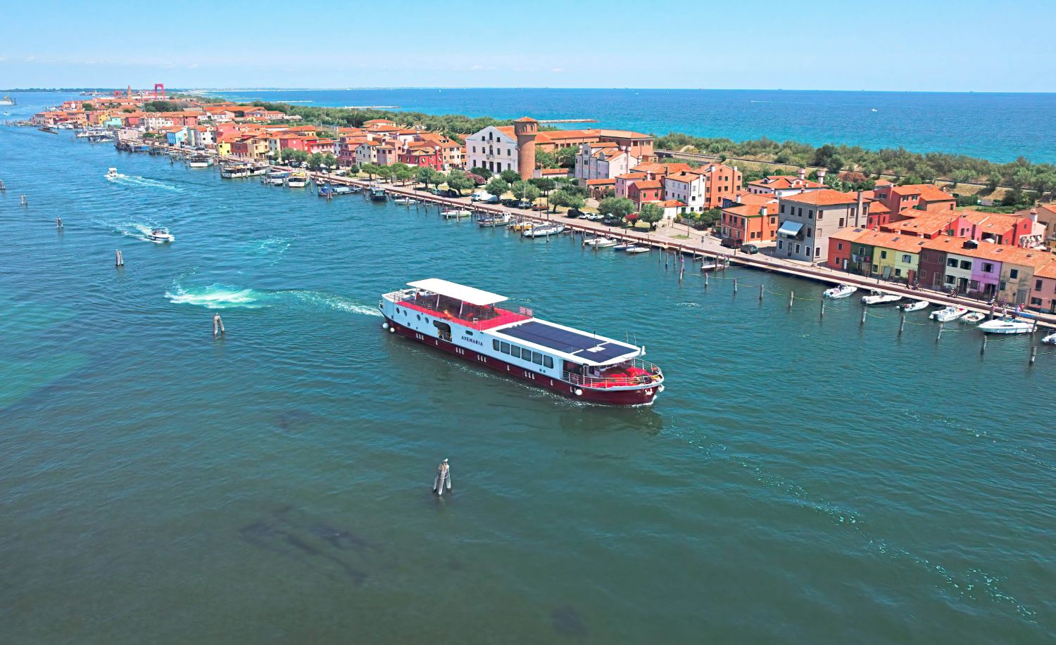 Das rot-weiße Boot Girolibero fährt auf der venezianischen Lagune mit einem Dorf aus bunten Häusern im Hintergrund