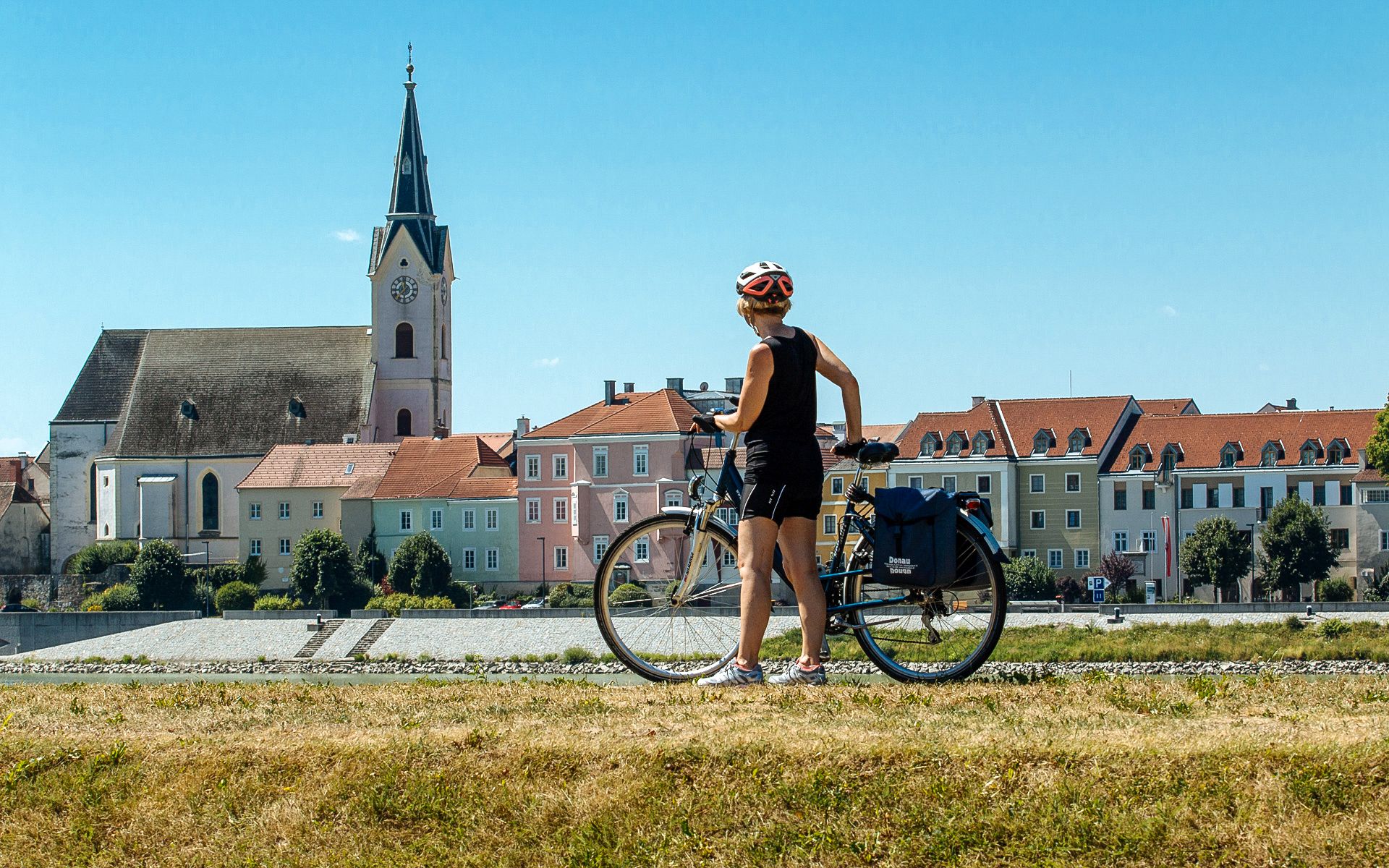 Ciclista con caschetto ferma ad osservare un paesino lungo la ciclabile del Danubio, chiesa con campanile, Austria