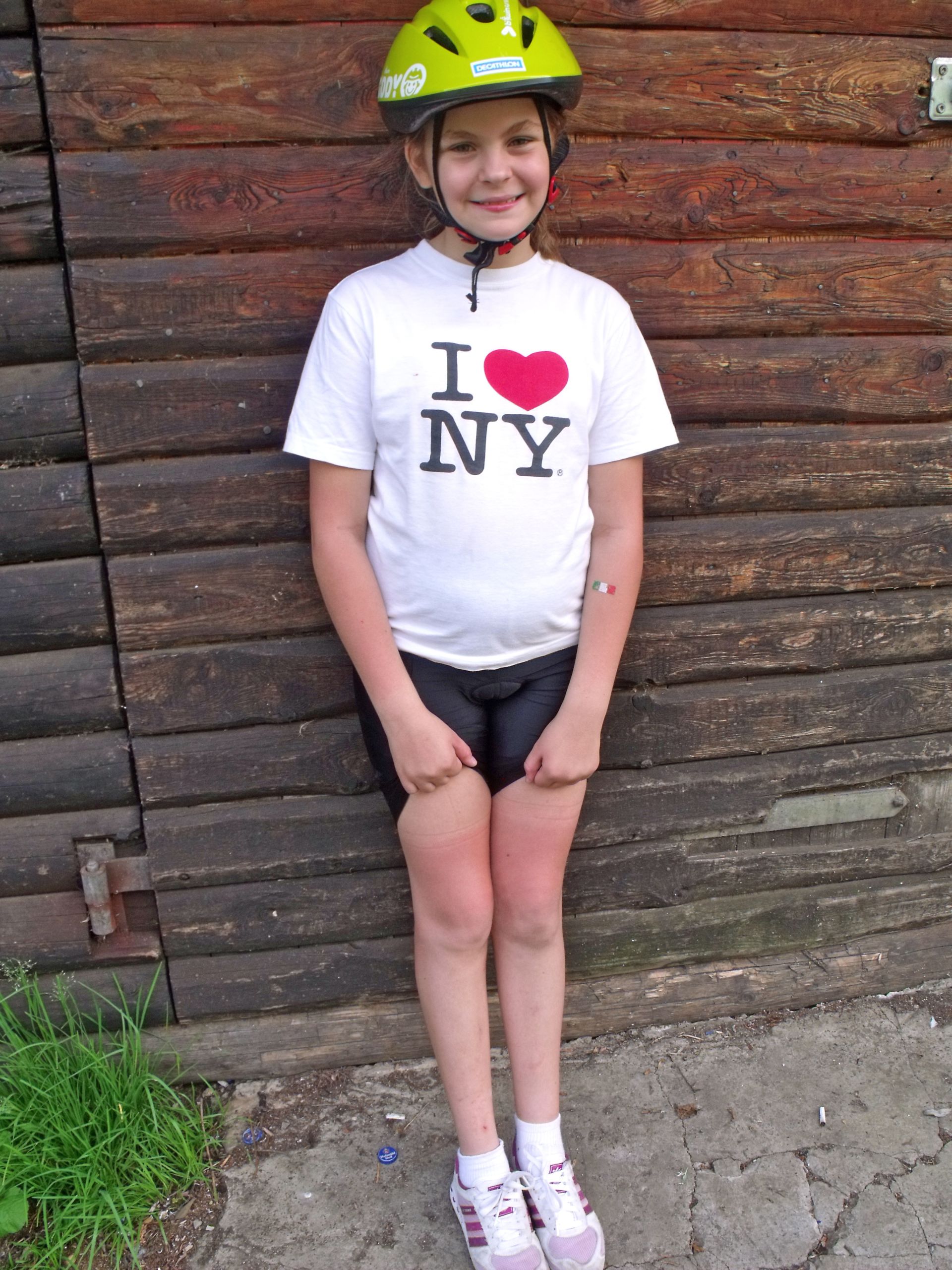 Bambina sorridente con casco appoggiata a una parete in legno, durante una vacanza in bici con Girolibero.