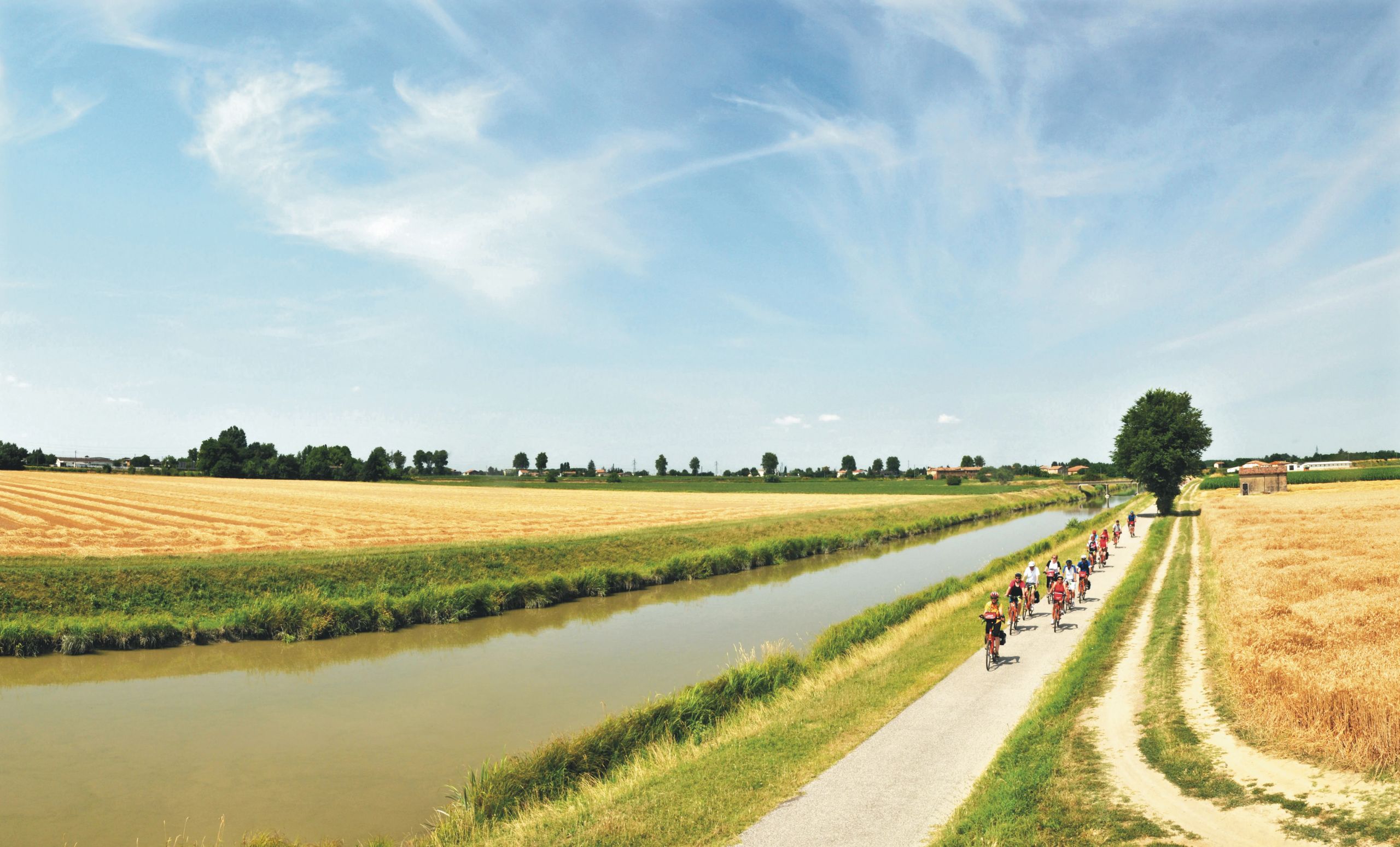 Radfahrer auf einem Radweg entlang eines Kanals, umgeben von Weizenfeldern und einem klaren Himmel, Italien