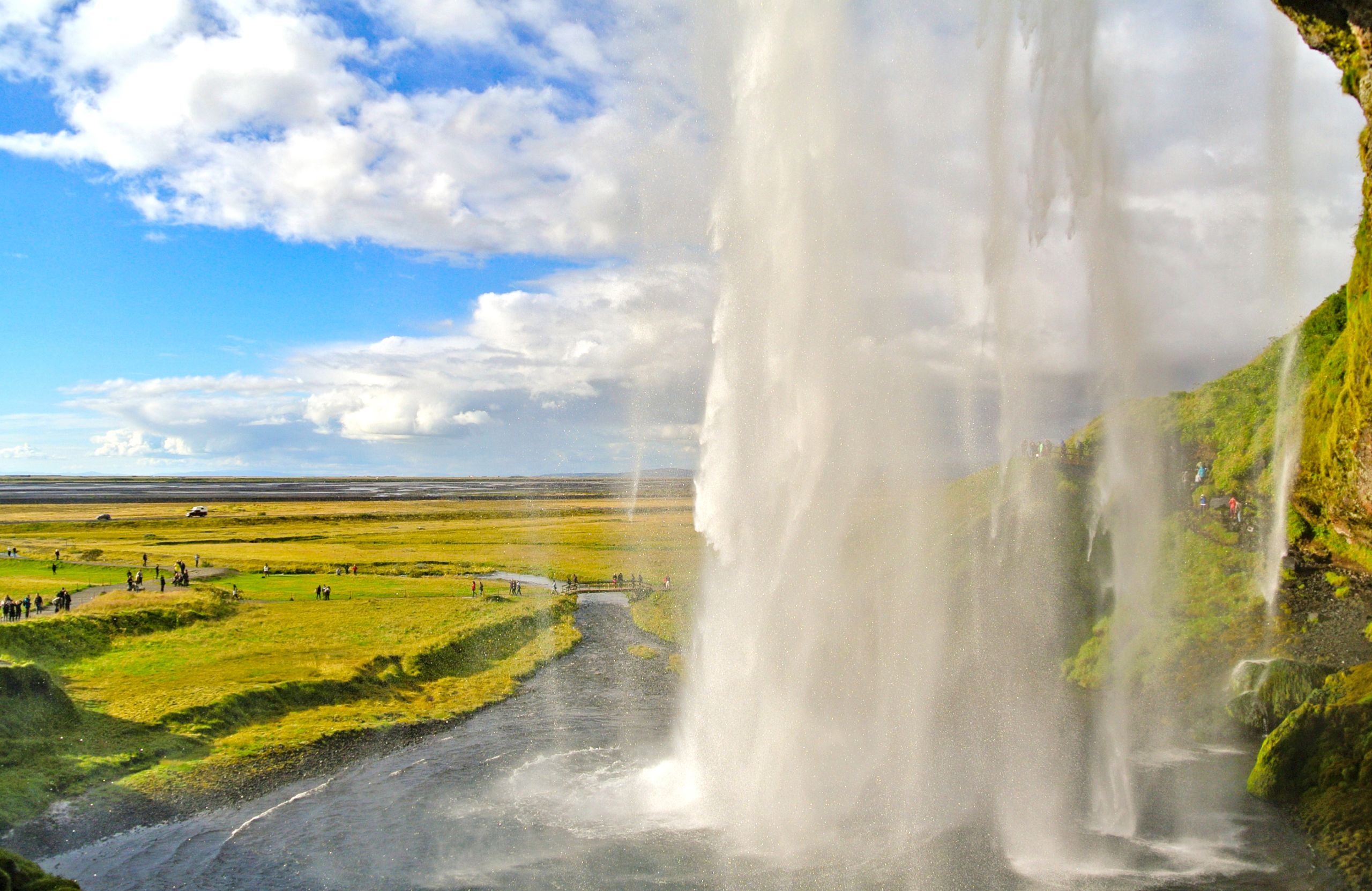 Vista da dietro della cascata Seljalandsfoss, con paesaggi verdi e cielo blu in Islanda.