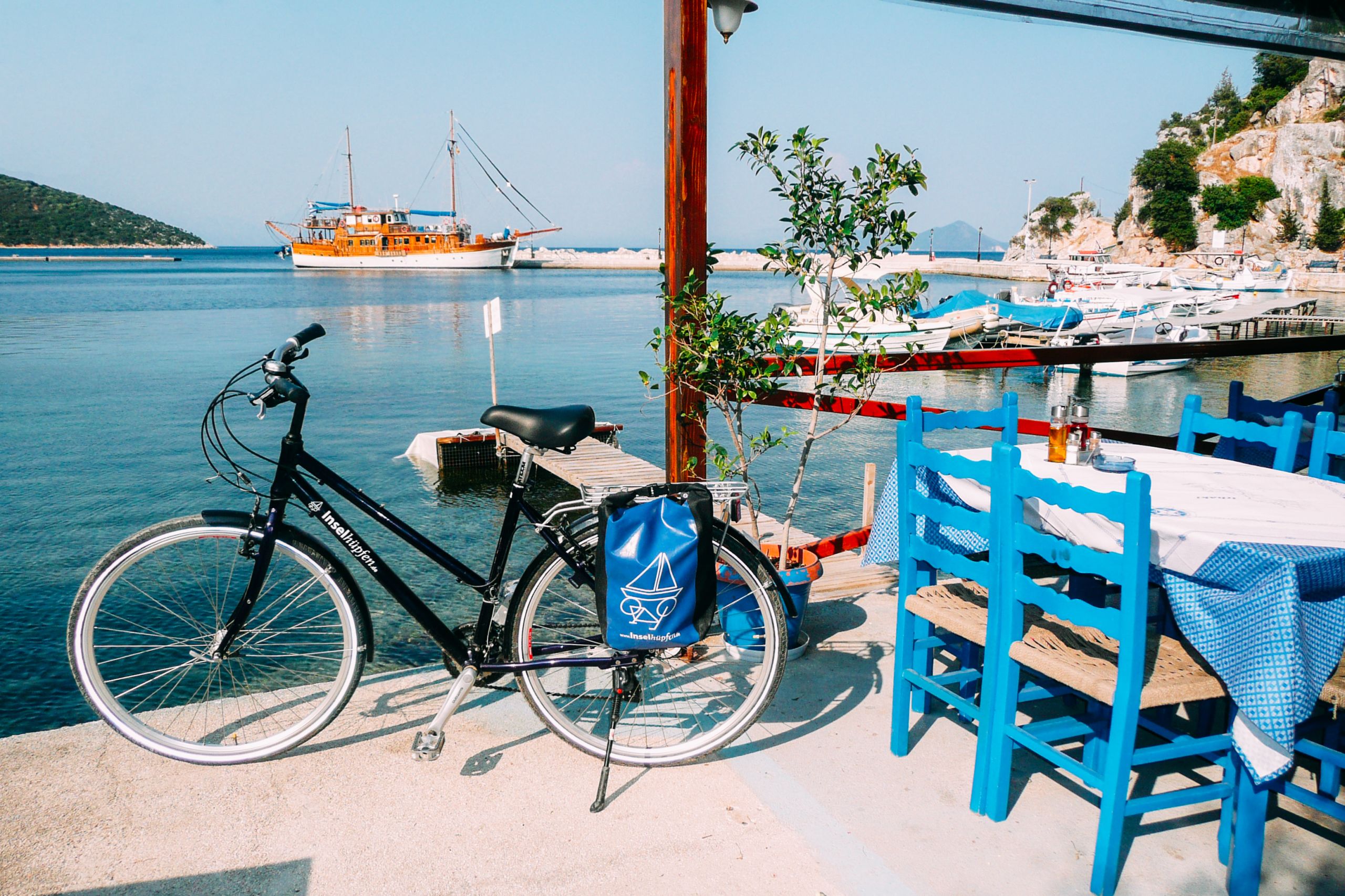 Bici vicino a tavolini e sedie blu in una terrazza con vista mare con barca in legno sullo sfondo, viaggi bici e barca 