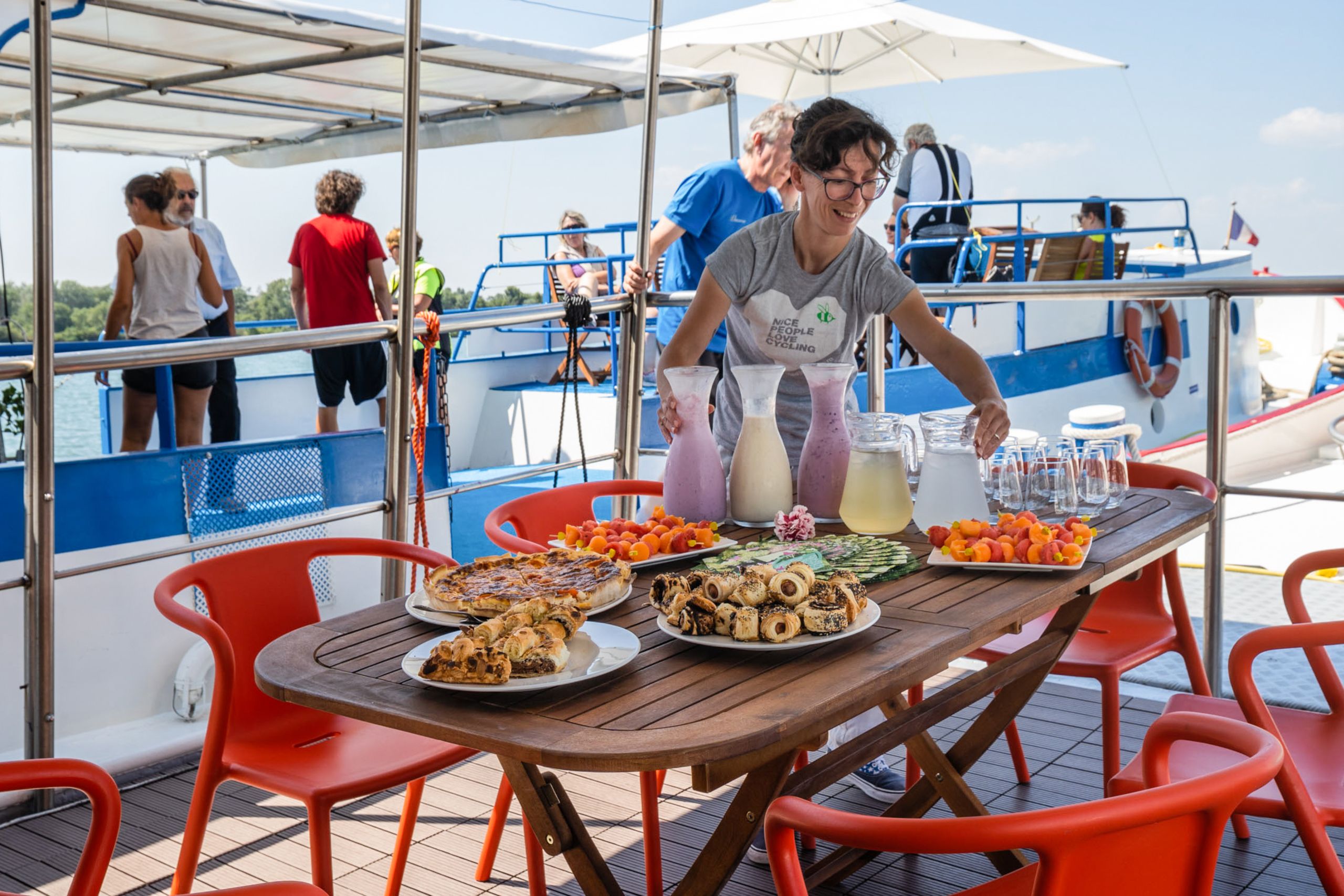Outdoor table with plates of local food on a boat, offering a panoramic view of the water, with a woman serving the meal
