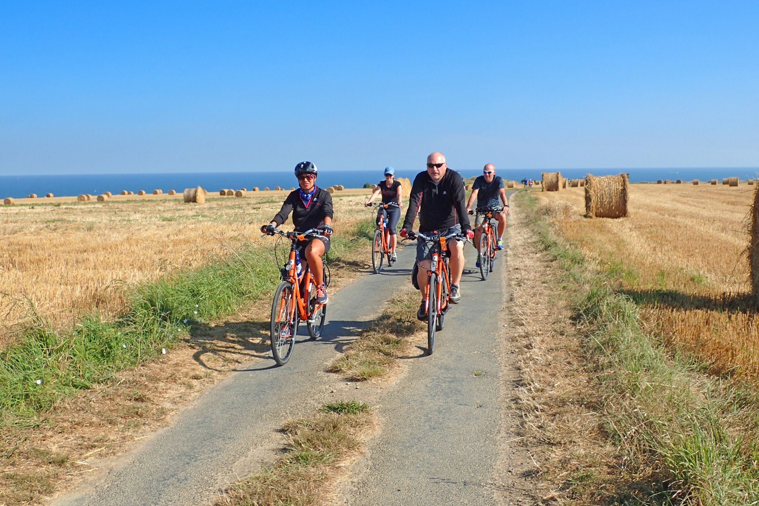 Gruppo di ciclisti in viaggio con Girolibero su un sentiero rurale in Bretagna, con balle di fieno sullo sfondo e cielo blu.