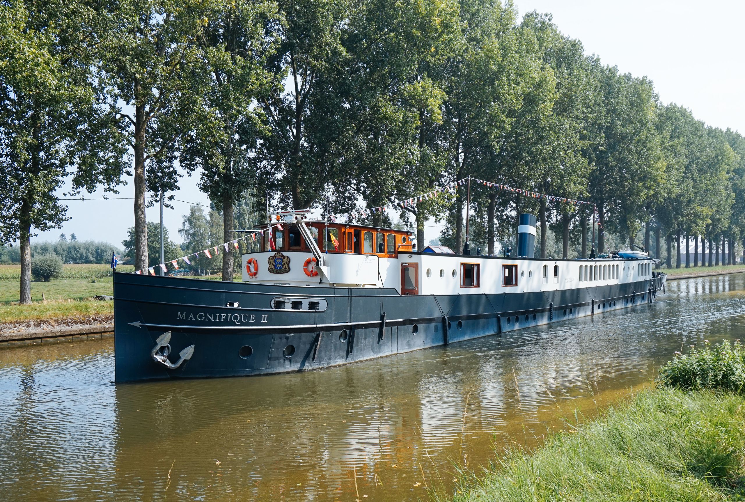 Wunderschönes Flussschiff entlang eines Kanals mit einer Reihe großer grüner Bäume am anderen Ufer