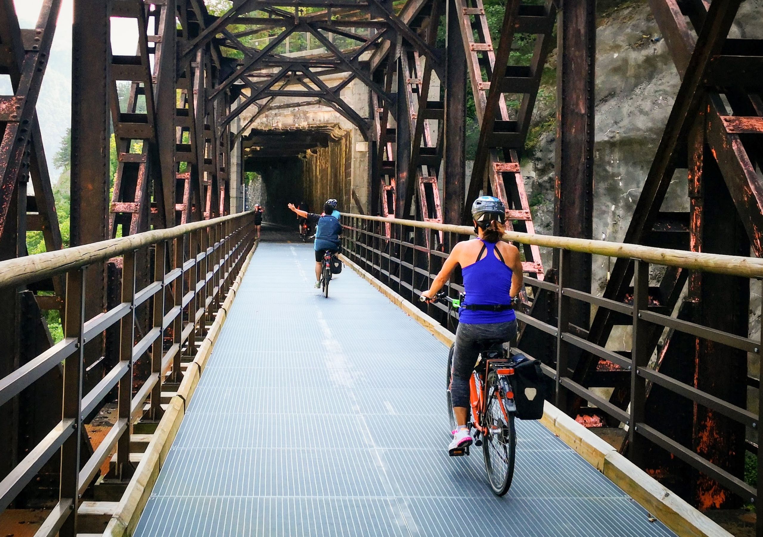 Ciclisti con casco e abbigliamento sportivo attraversano ponte sospeso in legno lungo la Ciclovia dell'Alpe Adria.