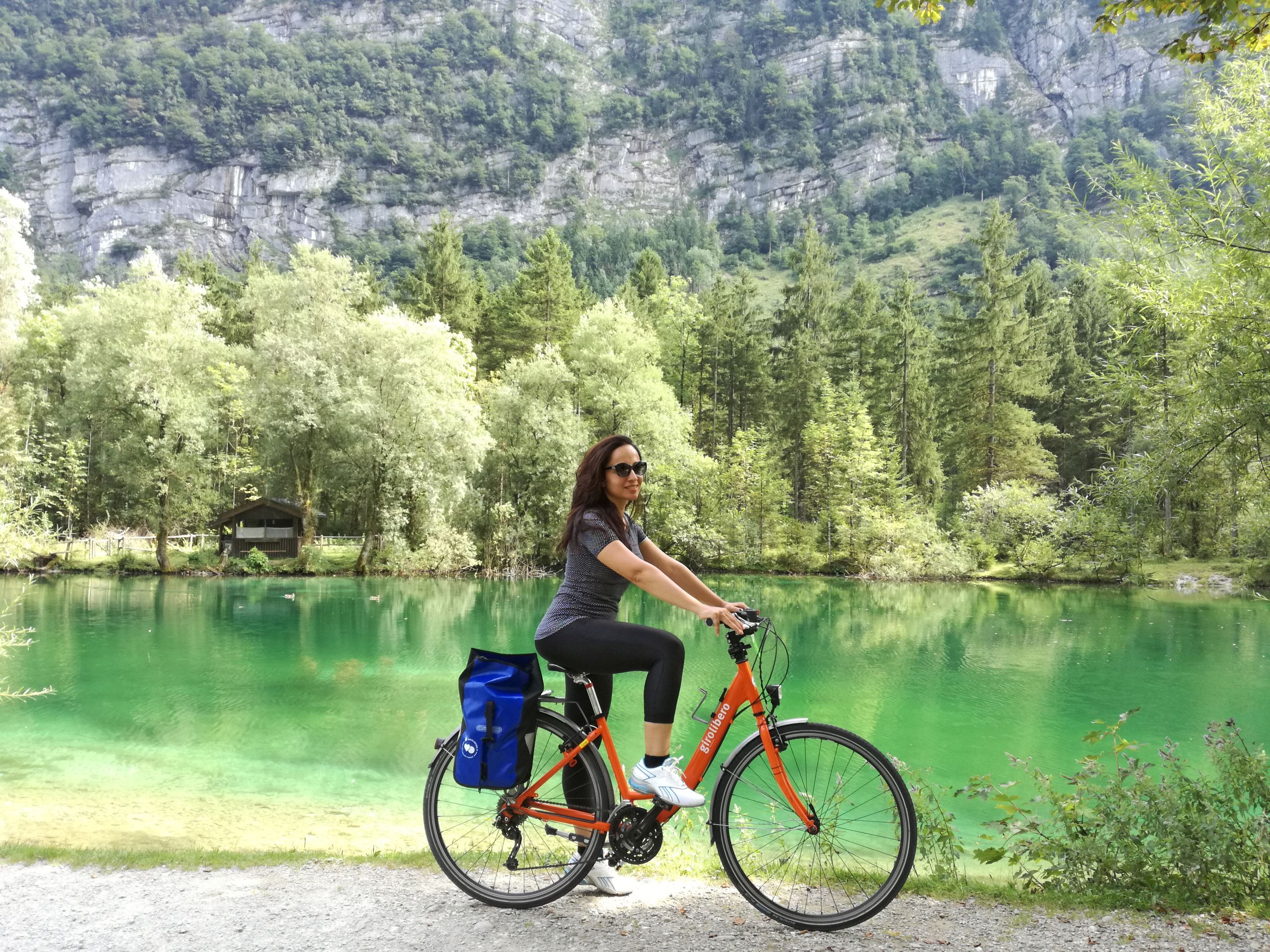 Ragazza in bici arancione "Girolibero" davanti a un laghetto alpino tra le montagne lungo la ciclabile dei Tauri