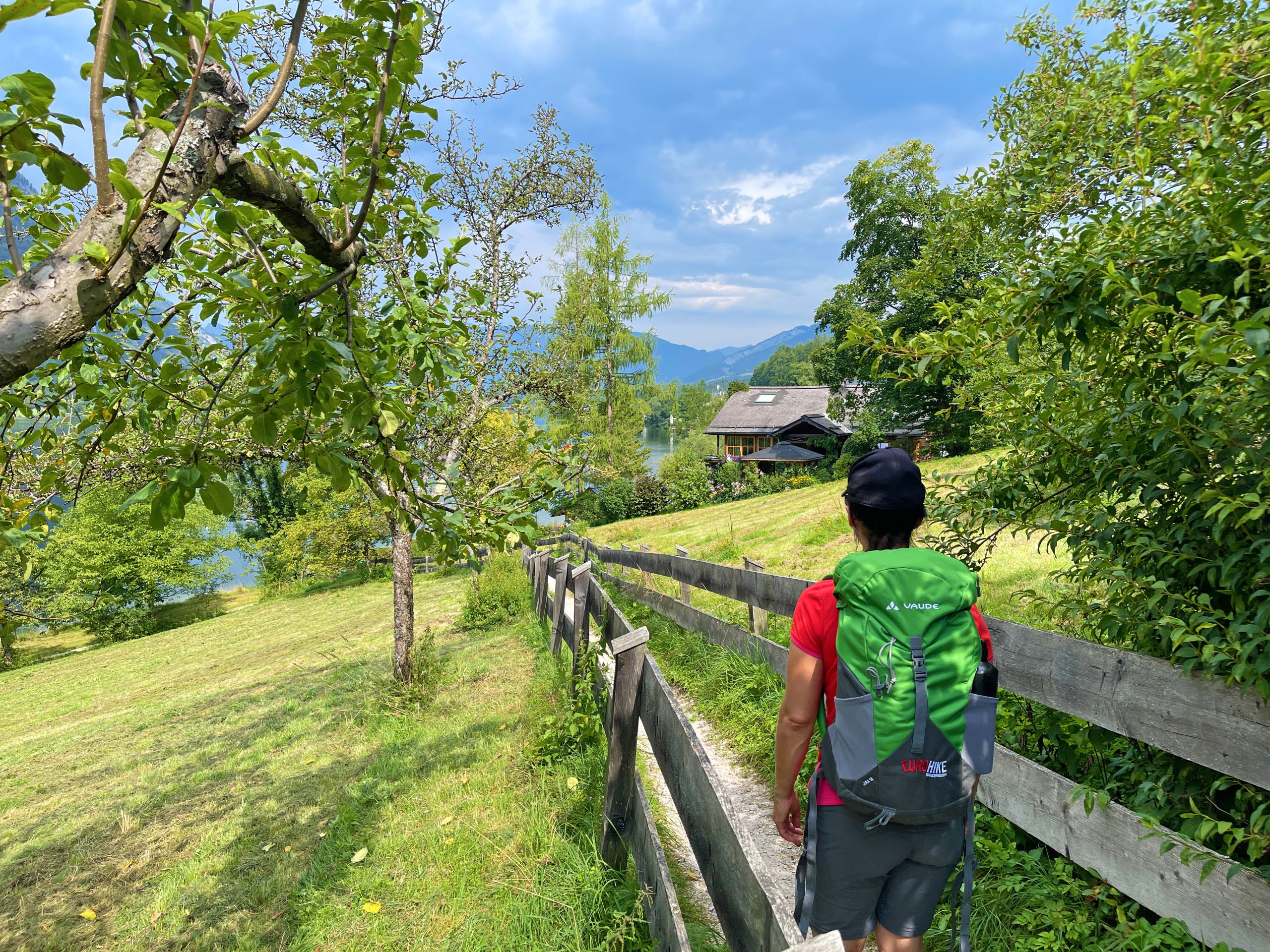 Escursionista cammina lungo una recinzione in legno con vista sul lago e montagne del Salzkammergut, trekking in Austria.