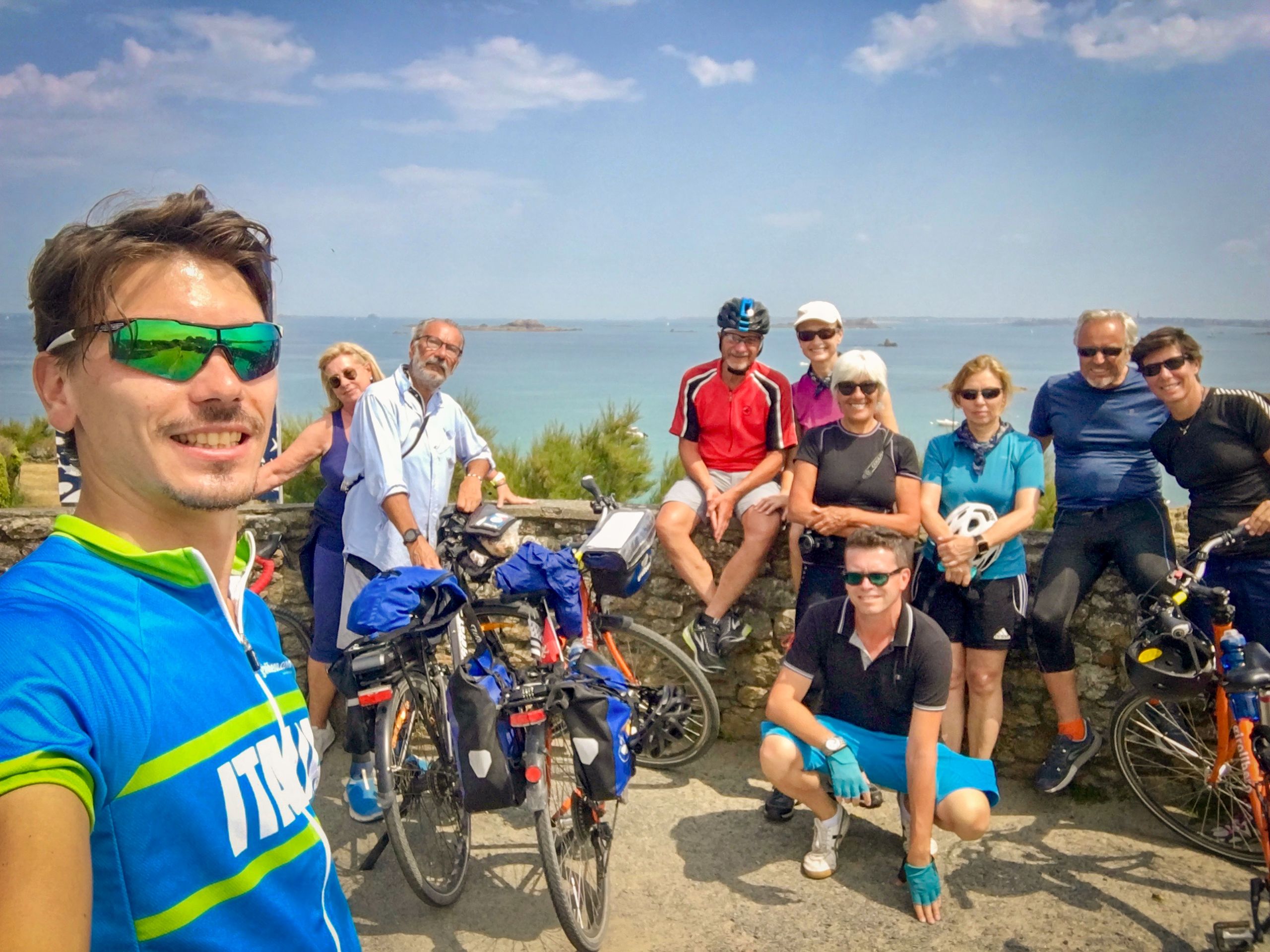 Gruppo di ciclisti in posa durante una pausa, con biciclette parcheggiate e paesaggio bretone sullo sfondo.Viaggi di gruppo in bici organizzati.
