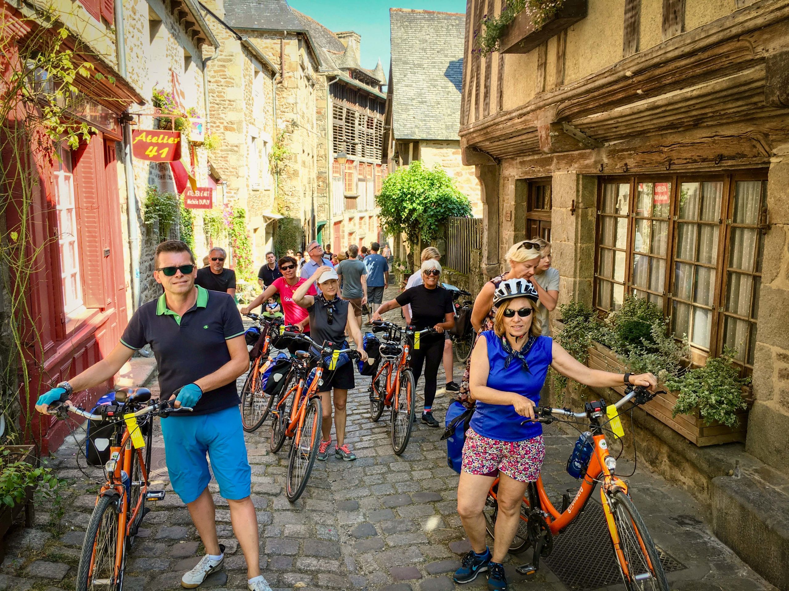 Ciclisti che percorrono le strade acciottolate di una cittadina medievale in Bretagna, circondati da edifici storici. Vacanze in bicicletta con Girolibero.