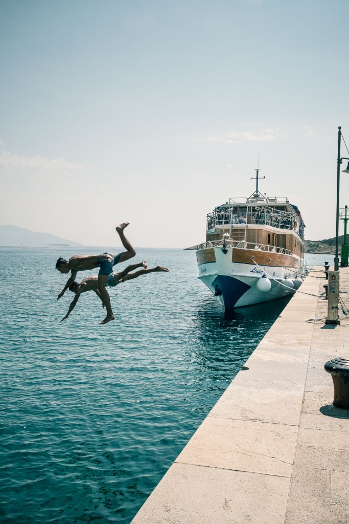 Two people diving into the sea from a pier with a docked boat, Croatia.