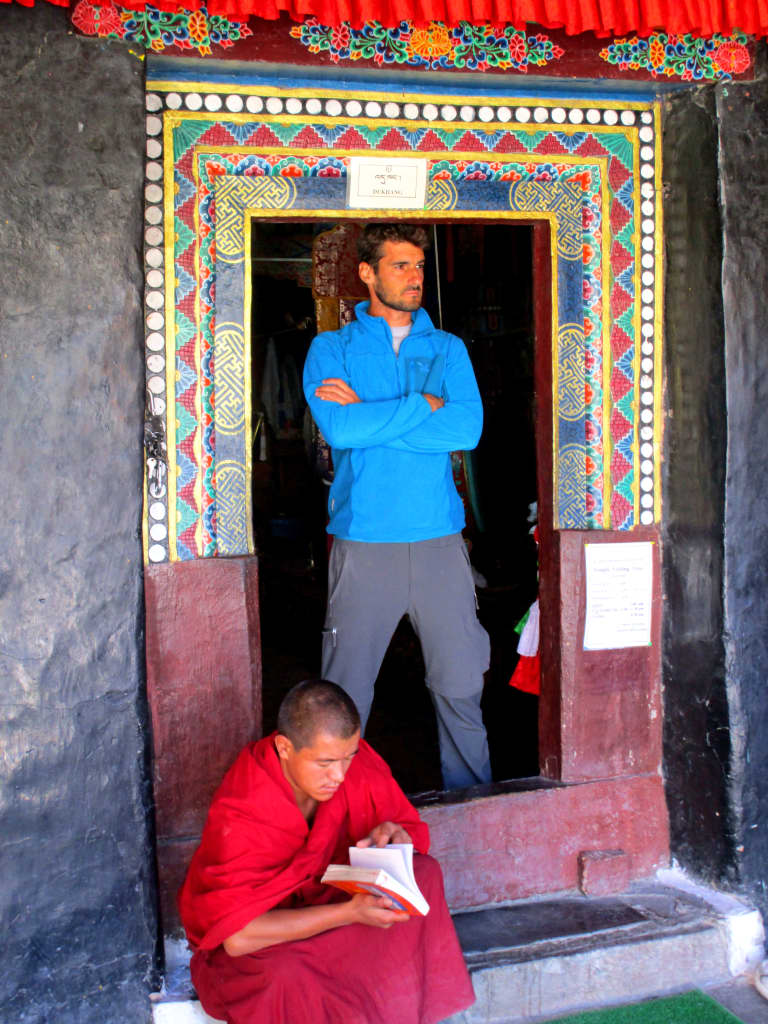 Portale dipinto del monastero Hemis in Ladakh, viaggiatore occidentale accanto a monaco tibetano assorto in lettura.