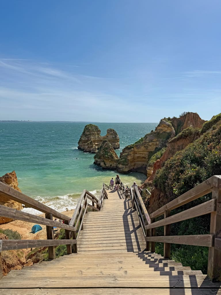 Scalinata panoramica in legno che scende verso la spiaggia di Praia do Camilo a Lagos, acque turchesi e faraglioni calcarei.