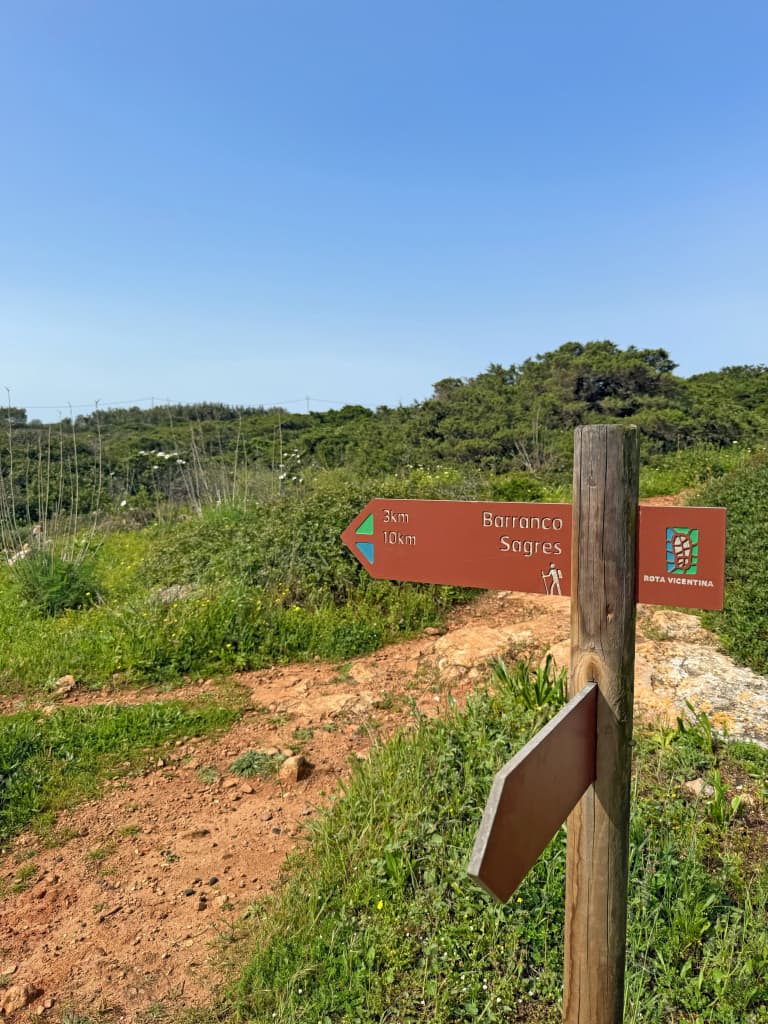 Segnaletica in legno della Rota Vicentina sullo sfondo verde del Parque Natural do Sudoeste Alentejano e Costa Vicentina.