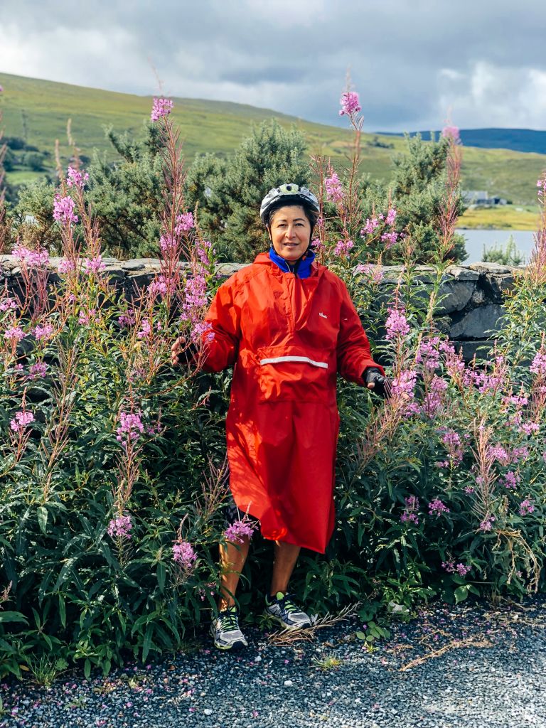 Viaggiatrice con caschetto e impermeabile rosso antipioggia in posa tra fiori selvatici lungo un sentiero nel Connemara.