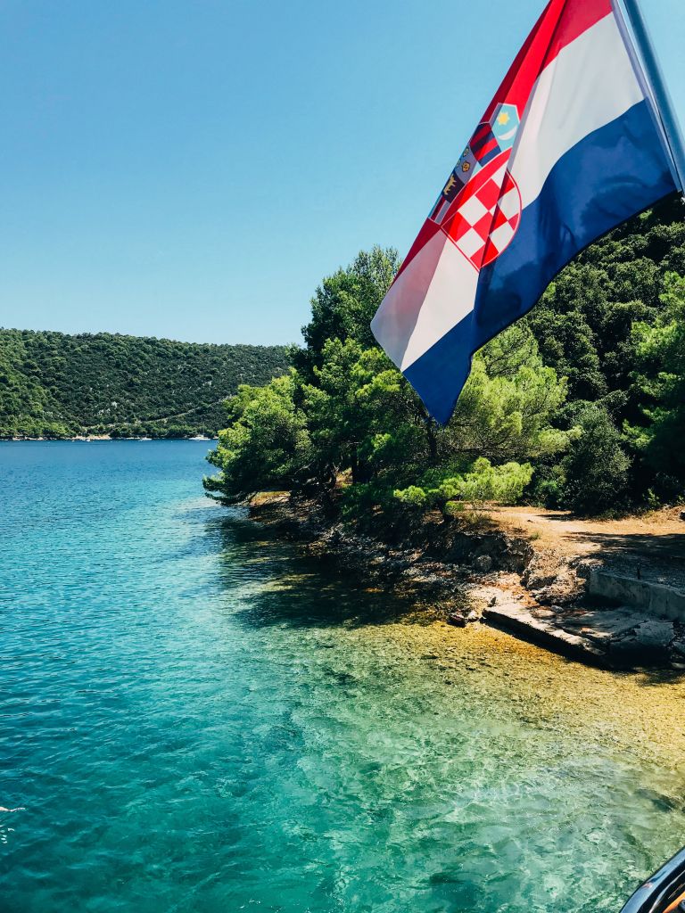 Felsige Küste mit Kiefern und kroatischer Flagge, die im Wind auf dem Deck eines Schiffes weht