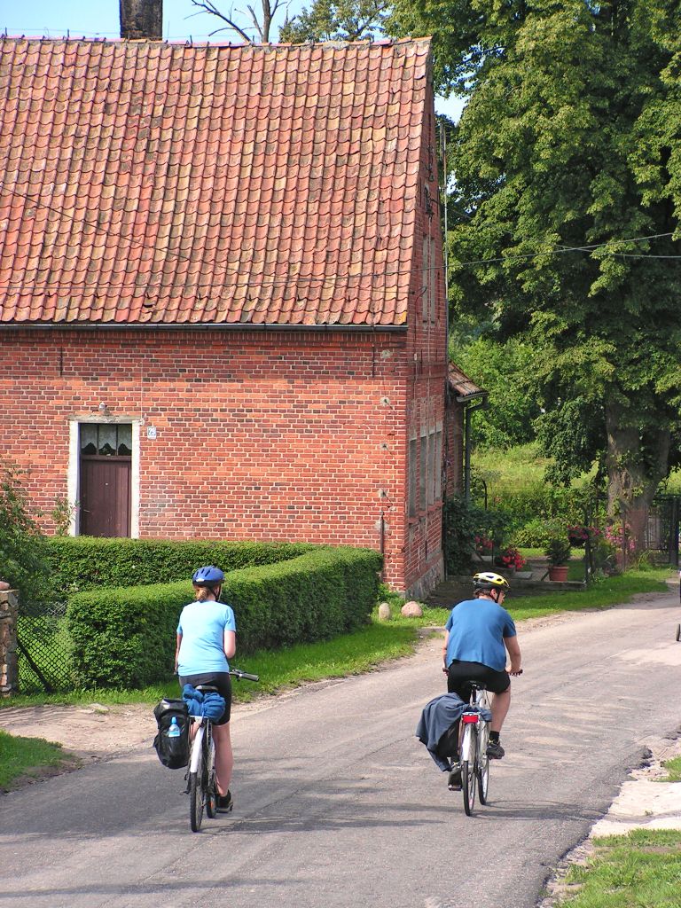 Due ciclisti in viaggio su una strada di campagna con case in mattoni tipiche dei Laghi Masuri, Polonia.