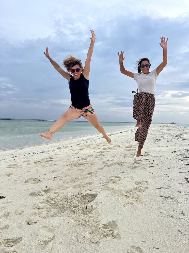Turiste in viaggio con Girolibero che saltano sulla spiaggia bianca di un’isola maldiviana, cielo sereno e mare sullo sfondo.