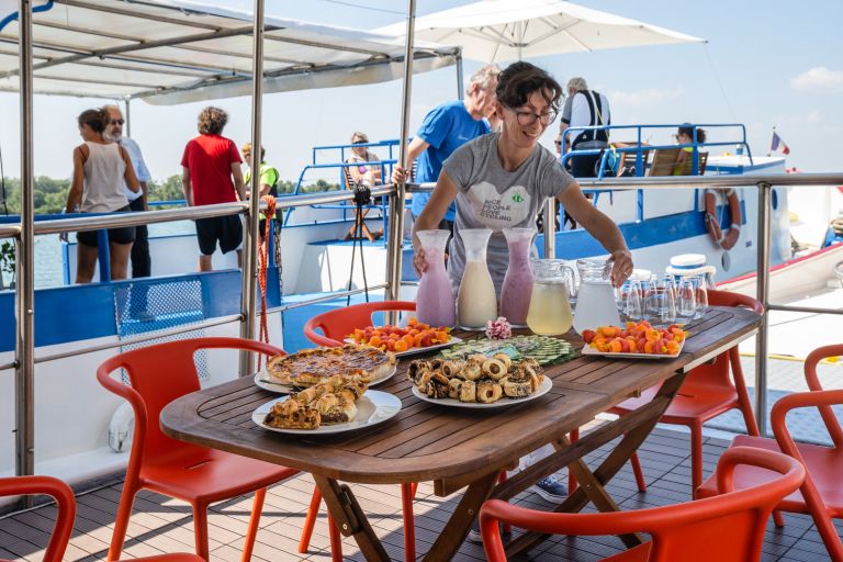 Outdoor table with plates of local food on a boat, offering a panoramic view of the water, with a woman serving the meal