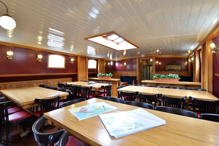 Spaziosa sala da pranzo della nave con tavoli in legno, mappa nautica aperta e soffitto illuminato.