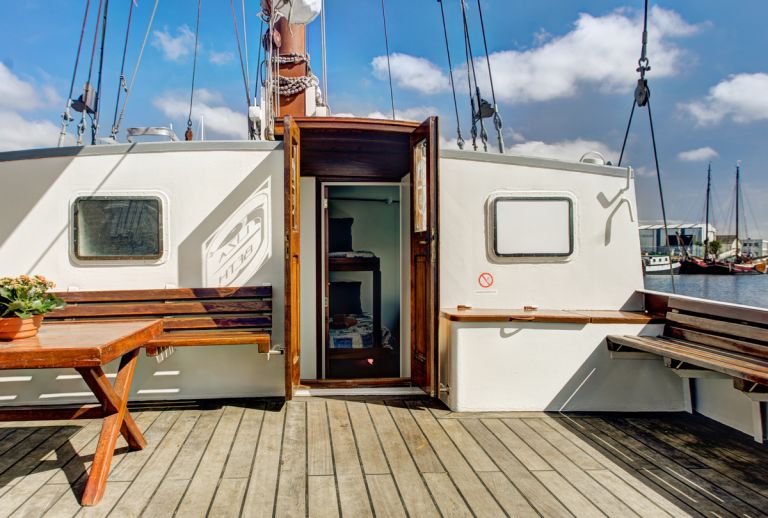 Ponte della nave con tavolino in legno, vista parziale delle vele e cielo sereno.