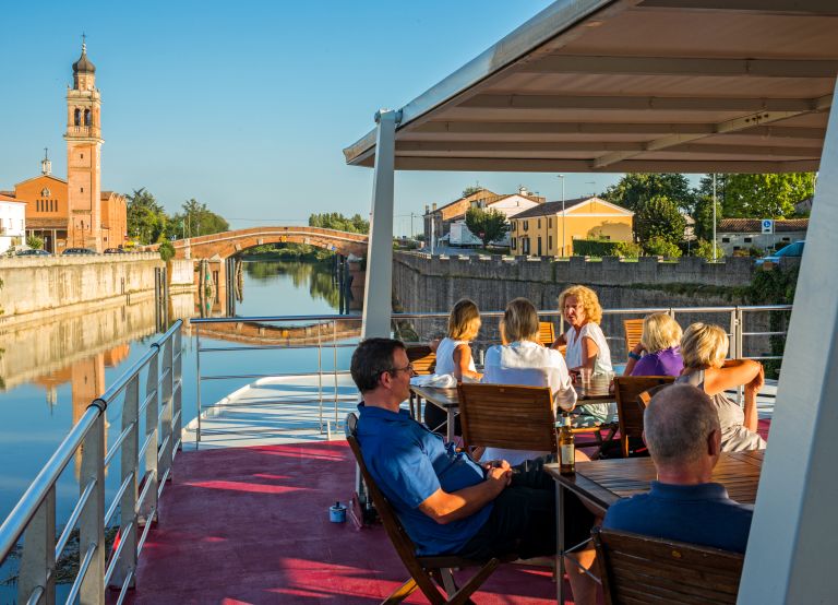 People dining outdoors on a riverboat with a view of Mantua, bike and boat tour, Italy.