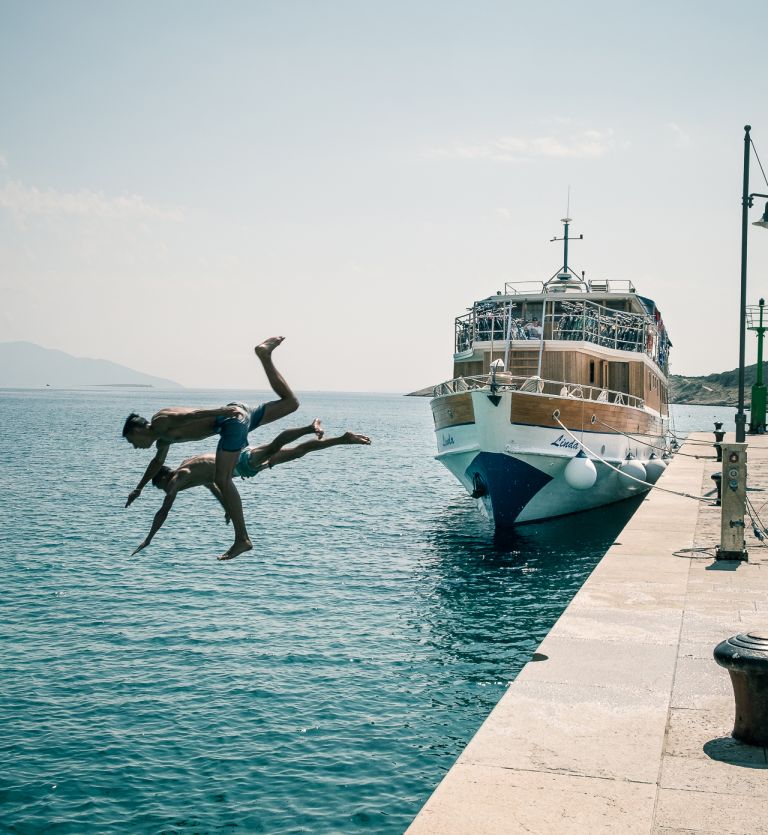 Two people diving into the sea from a pier with a docked boat, Croatia.