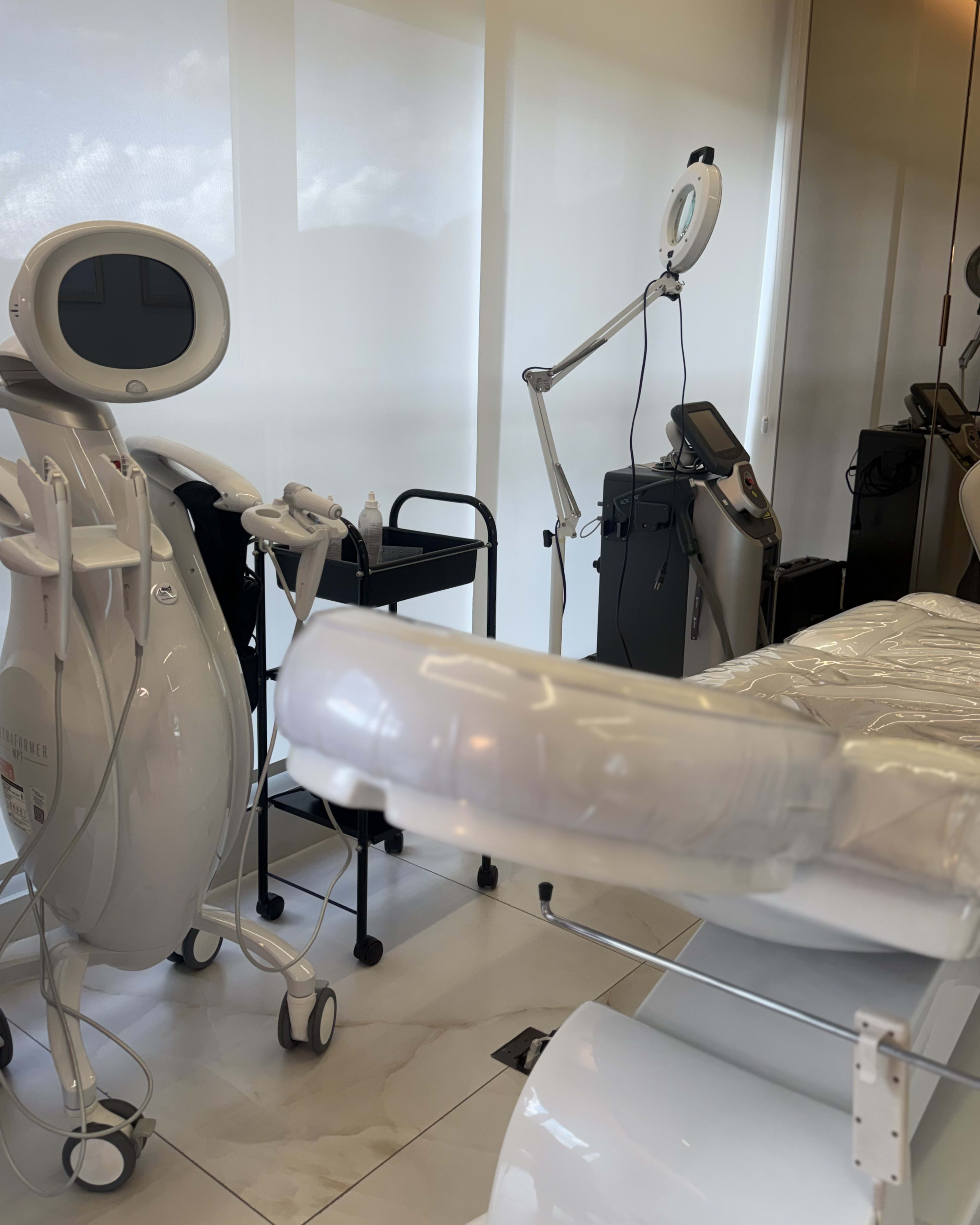 Sala de procedimento com maca preparada, lupa com iluminação e equipamentos de tecnologia estética