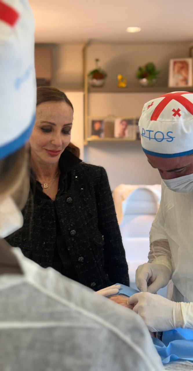 Dra. Gabriela Dawson acompanhando um procedimento realizado por profissionais paramentados com touca, avental e luvas