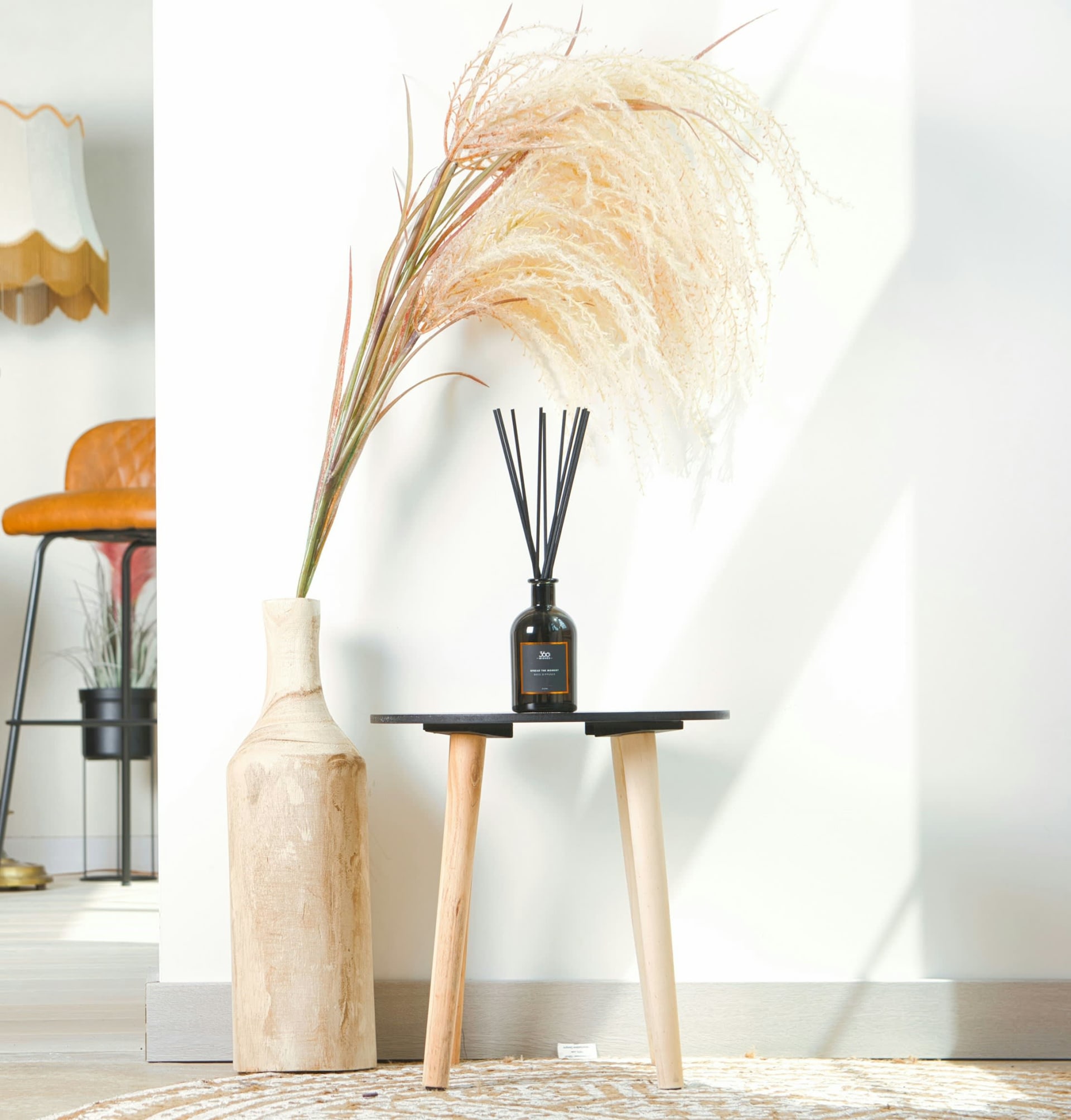 Dark glass reed diffuser on a small wooden side table beside a vase of pampas grass in a bright, airy room