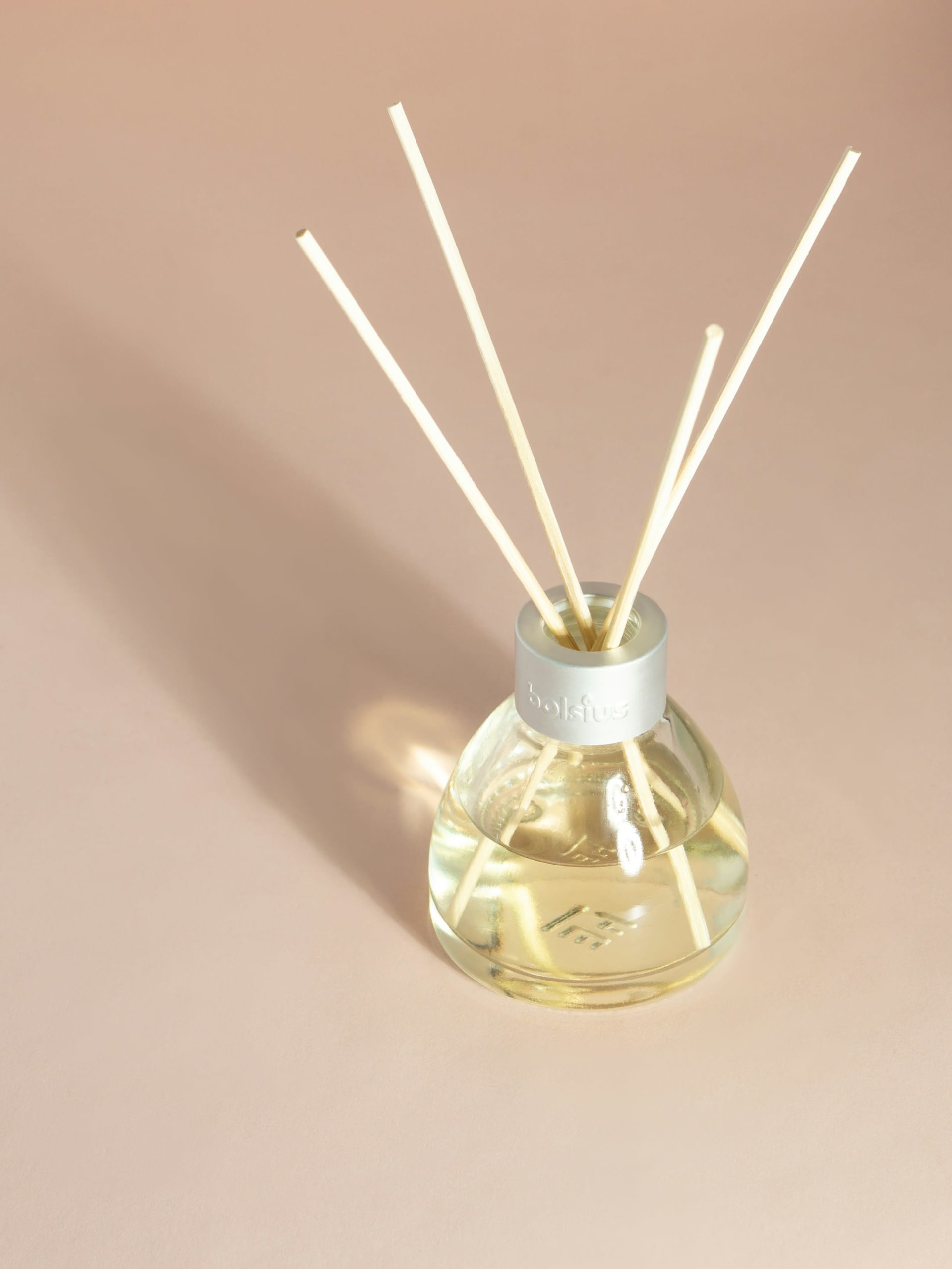 Clear glass reed diffuser with white reeds in bright light against a warm peach wall