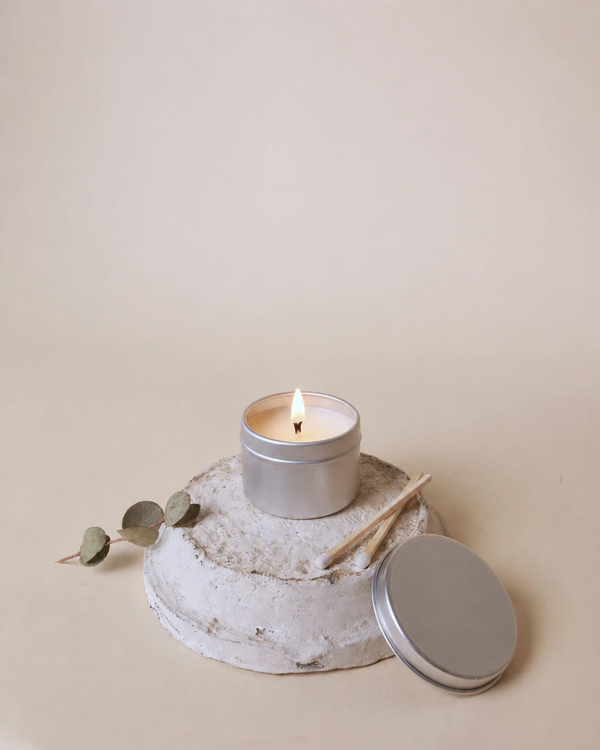 Lit cream candle in a tin on a stone plinth with eucalyptus against a beige backdrop
