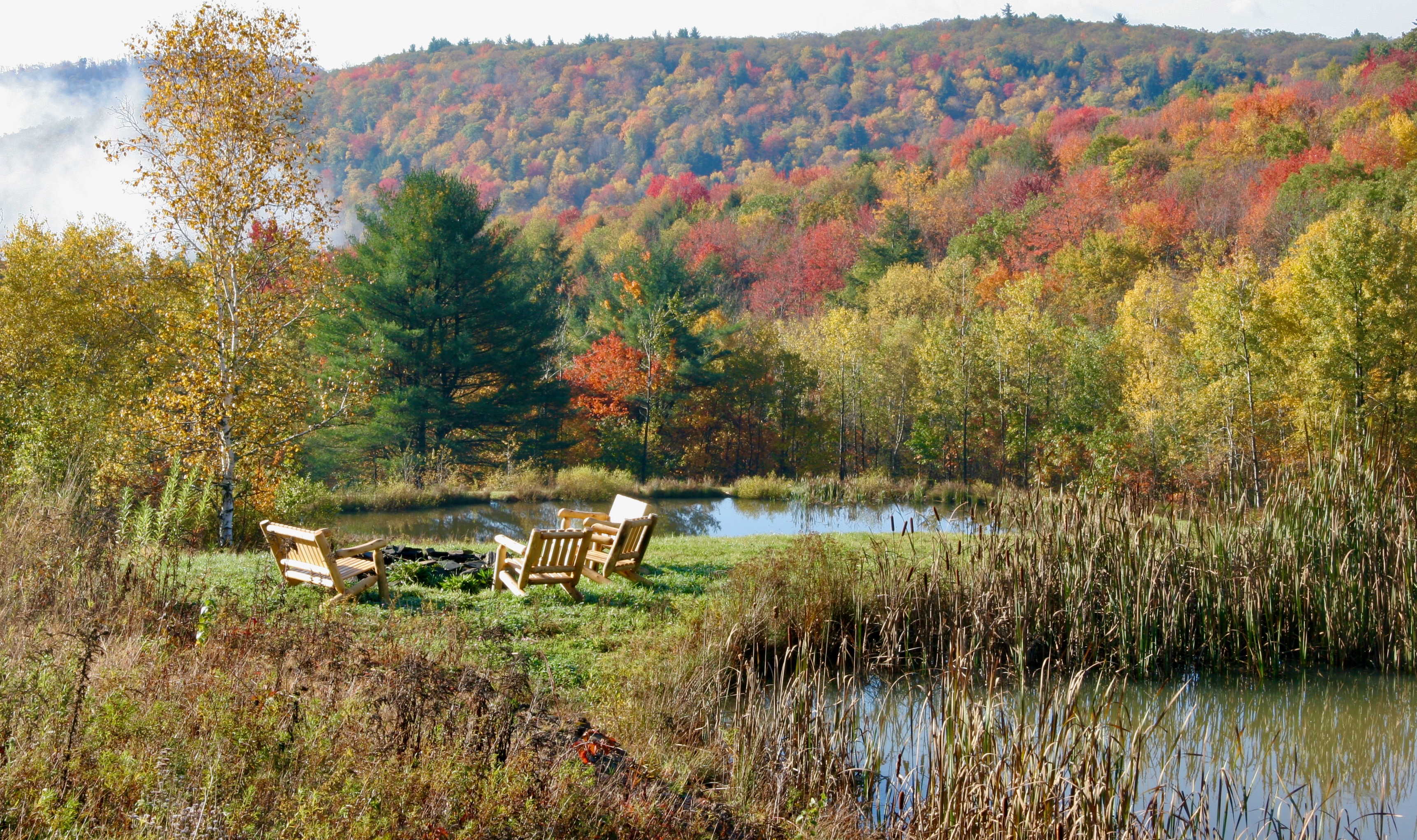Adirondack chairs at fire pit by pond with fall foliage mountains at Mountain Meadows private Catskills retreat